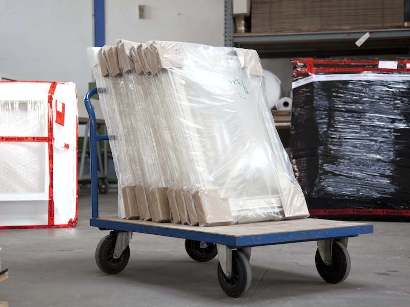 Rollwagen in der Halle mit folienverpackten Elementen und Kantenschutz aus Pappe, daneben Gitterboxen.