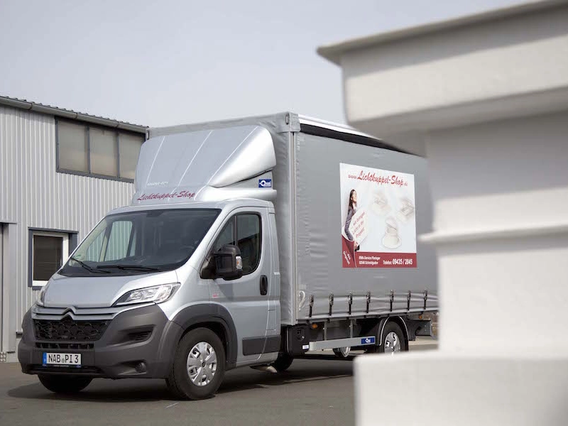 Silberner Transporter vor der Halle, auf der Plane die Werbung des Lichtkuppel-Shops mit Telefonnummer.