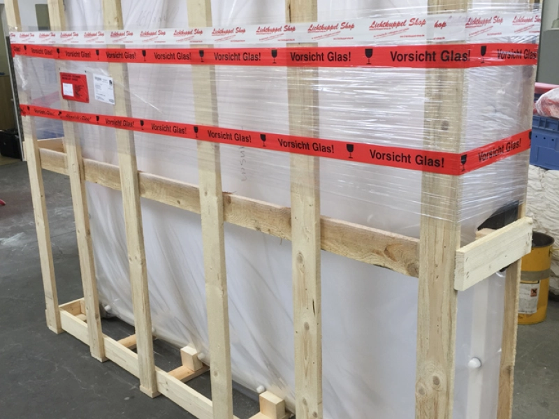 Hohe Transportkiste aus Holzlatten mit folienverpacktem Inhalt, umlaufend rotes Klebeband mit der Aufschrift &bdquo;Vorsicht Glas!&ldquo;.
