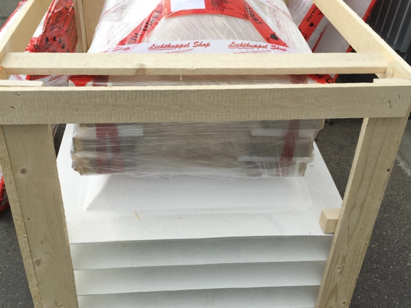 Holzverschlag als Transportkiste: oben eine folienverpackte Lichtkuppel mit rotem Klebeband, darunter gestapelte Aufsatzkr&auml;nze.
