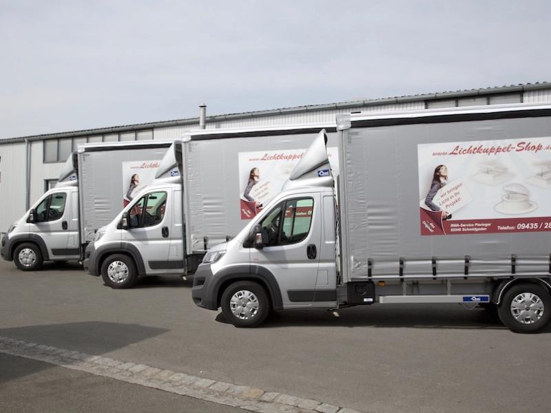 Drei silberne Transporter mit Planenaufbau und Lichtkuppel-Shop-Beschriftung stehen hintereinander vor der Halle.