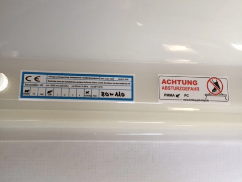 CE-Etikett und Warnaufkleber &bdquo;Achtung Absturzgefahr&ldquo; nebeneinander auf dem Rand einer Lichtkuppel.