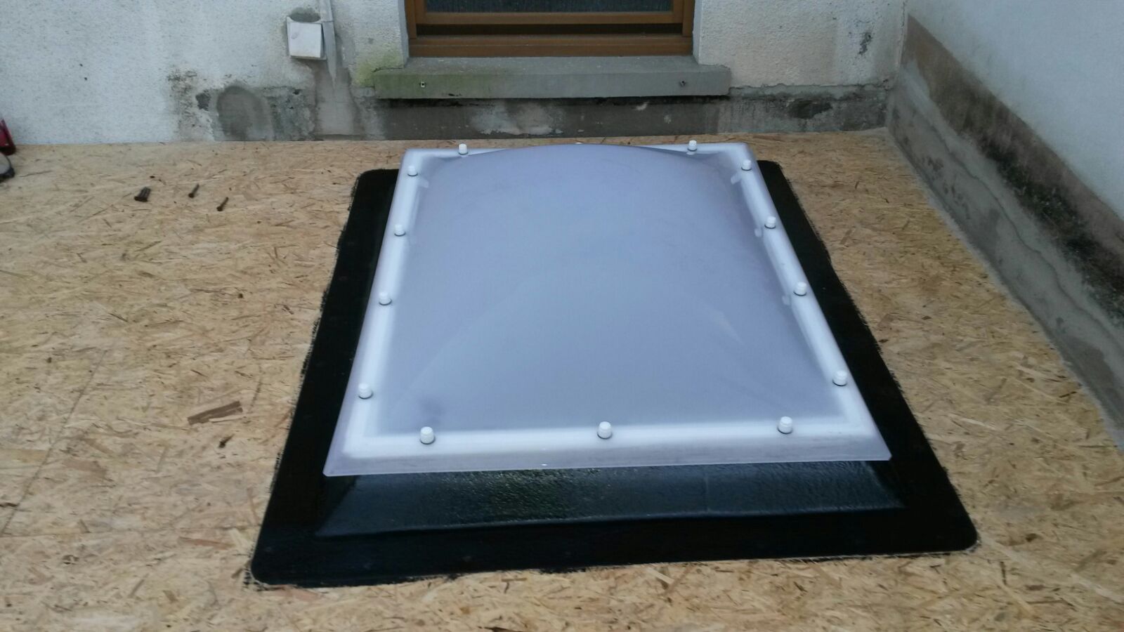 Lichtkuppel auf einem schwarz eingedichteten Aufsatzkranz, davor OSB-Platten, im Hintergrund eine Hauswand.