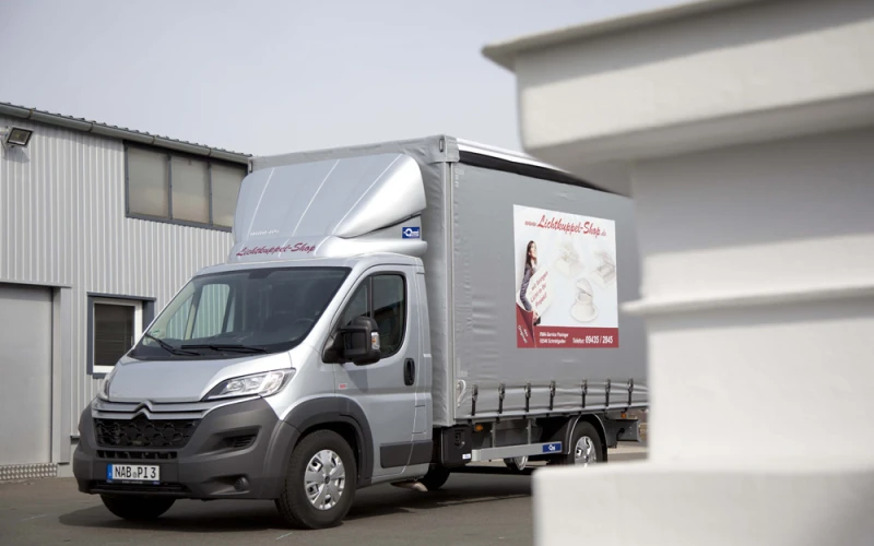 Silberner Lkw mit Planenaufbau und der Beschriftung Lichtkuppel-Shop, abgestellt vor einer Halle.