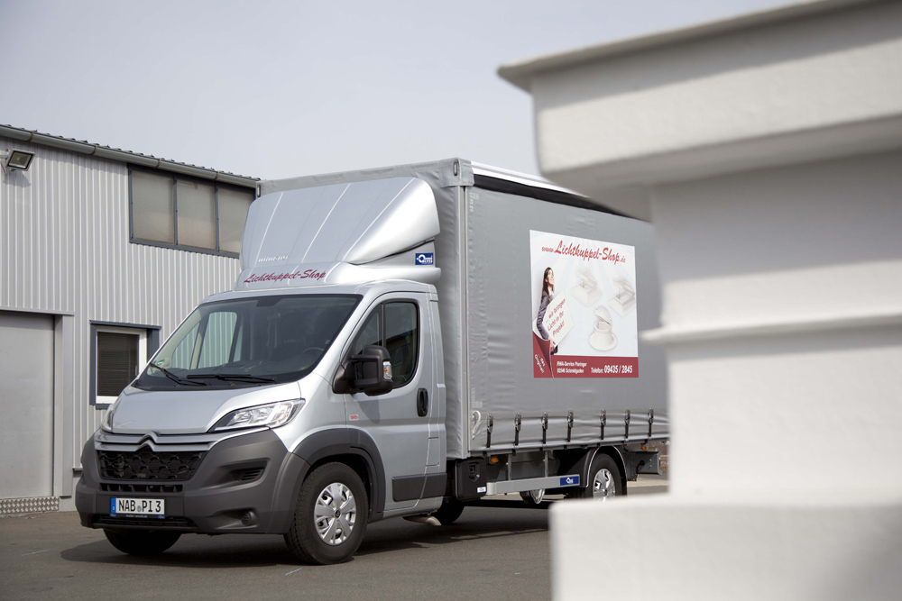 Silberner Lkw mit Planenaufbau und der Beschriftung Lichtkuppel-Shop, abgestellt vor einer Halle.