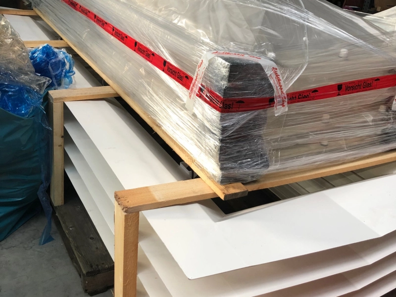 Langes, mit Folie und rotem Vorsicht-Glas-Band gesichertes Paket auf einem Holzgestell in der Halle, darunter gestapelte wei&szlig;e Platten.