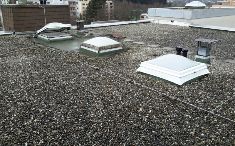 Kiesbedecktes Flachdach mit mehreren Lichtkuppeln unterschiedlicher Bauart, im Hintergrund Wohnh&auml;user.