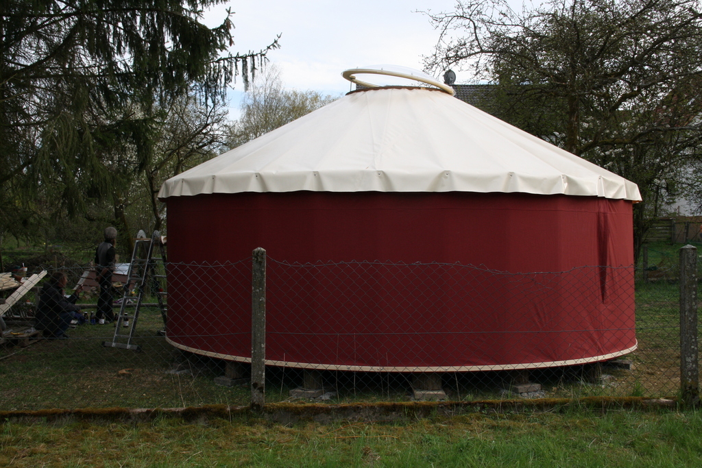 Jurte mit roter Wandbespannung und hellem Dach mit runder Lichtkuppel, davor ein Garten mit Bäumen und zwei Personen.