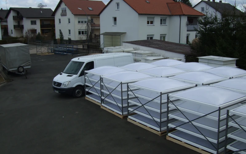 Hof mit Palettengestellen voller gestapelter Lichtkuppeln, daneben ein Transporter, im Hintergrund Wohnh&auml;user.