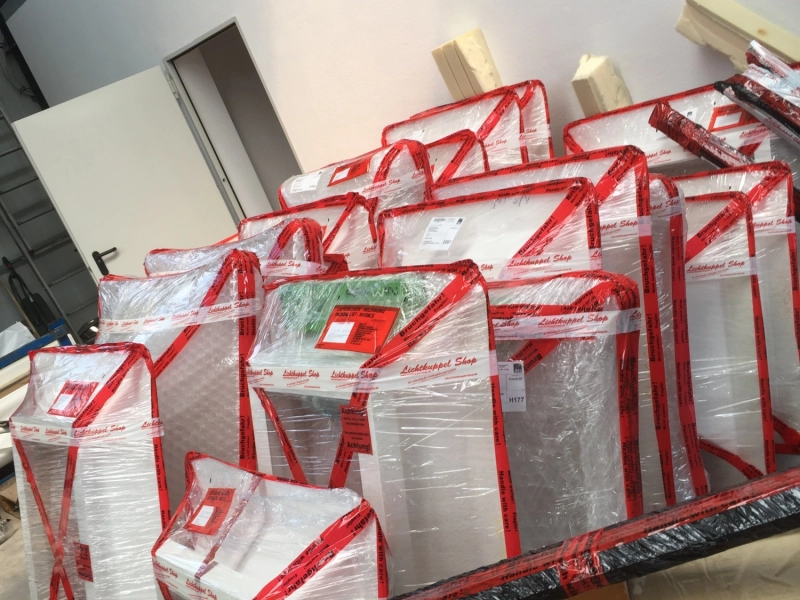 Mehrere in Folie und rotes Lichtkuppel-Shop-Band verpackte Lichtkuppeln, schr&auml;g aneinandergestellt, im Vordergrund ein Packtisch mit Schere und Folienrolle.