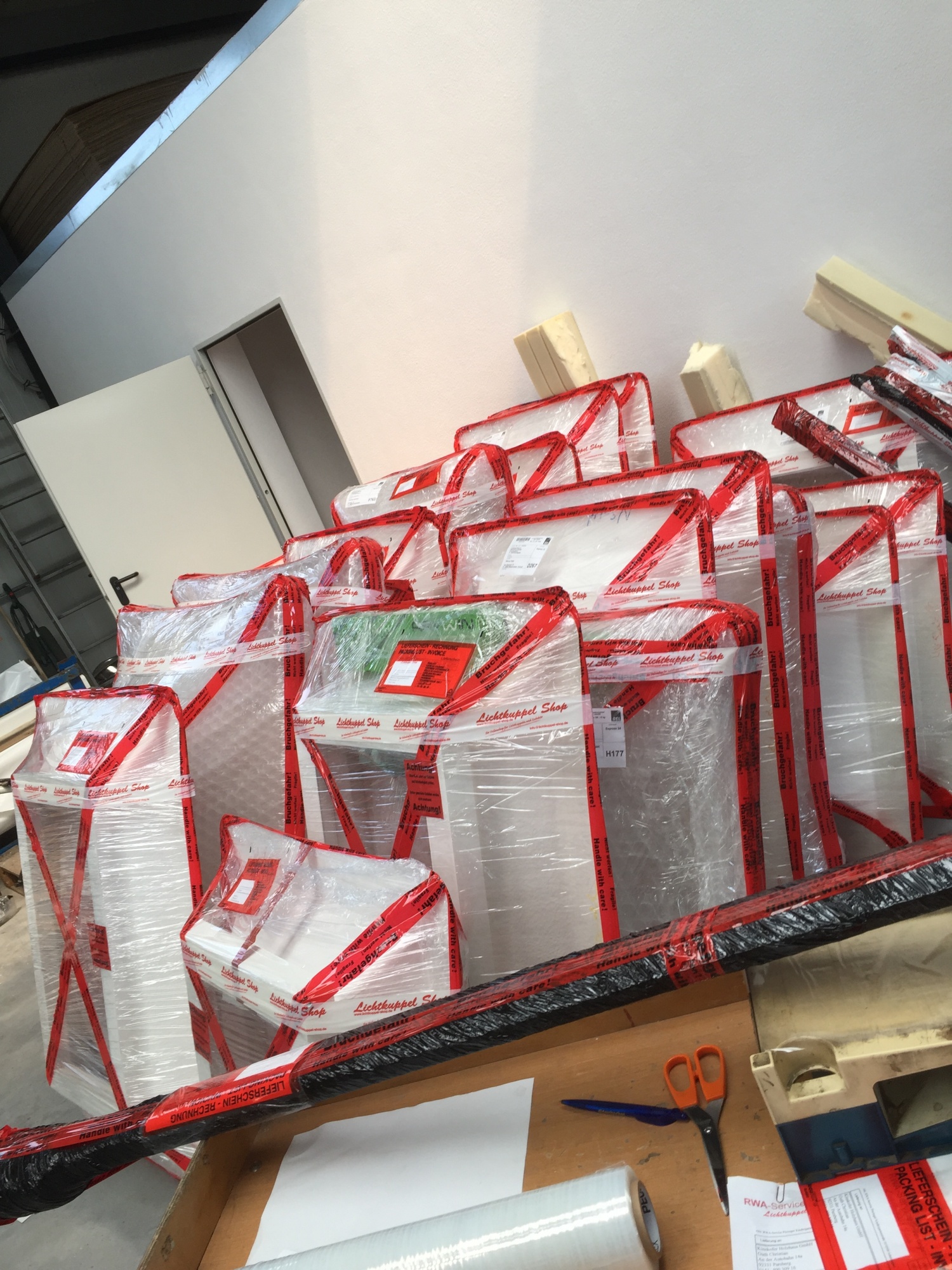 Mehrere in Folie und rotes Lichtkuppel-Shop-Band verpackte Lichtkuppeln, schräg aneinandergestellt, im Vordergrund ein Packtisch mit Schere und Folienrolle.