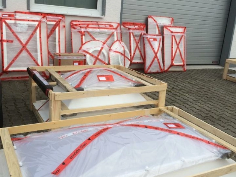 Verpackte Lichtkuppeln in flachen Holzrahmen auf dem Hof, dahinter an die Hauswand gelehnte, rot verschn&uuml;rte Pakete.