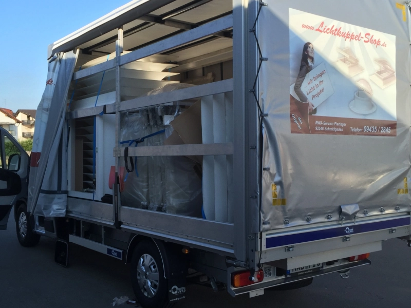 Lastwagen mit ge&ouml;ffneter Plane, darin verpackte Lichtkuppeln und Aufsatzkr&auml;nze, auf der Plane die Aufschrift www.Lichtkuppel-Shop.de, RWA-Service Pieringer, 92546 Schmidgaden.