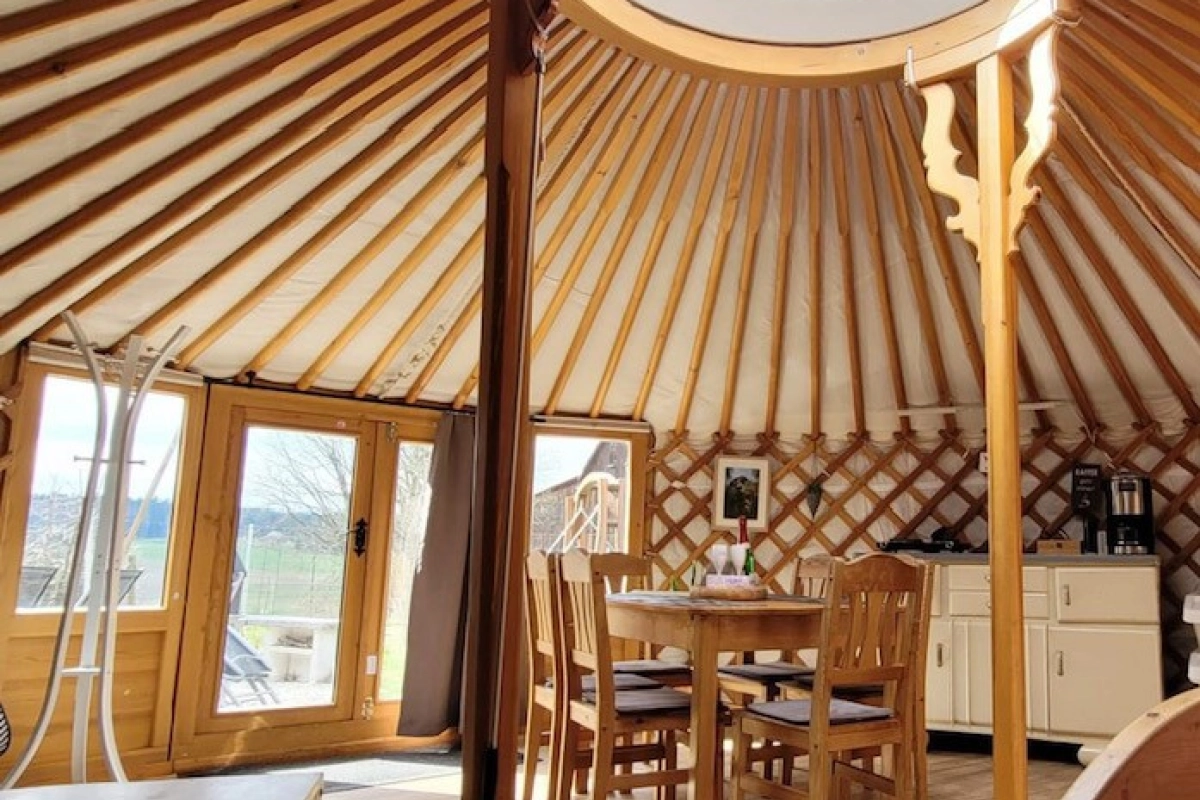 Blick in eine eingerichtete Jurte: Holzgitterwand, Speichendach und in der Mitte die runde Lichtkuppel, darunter Esstisch, K&uuml;chenzeile und Bett.