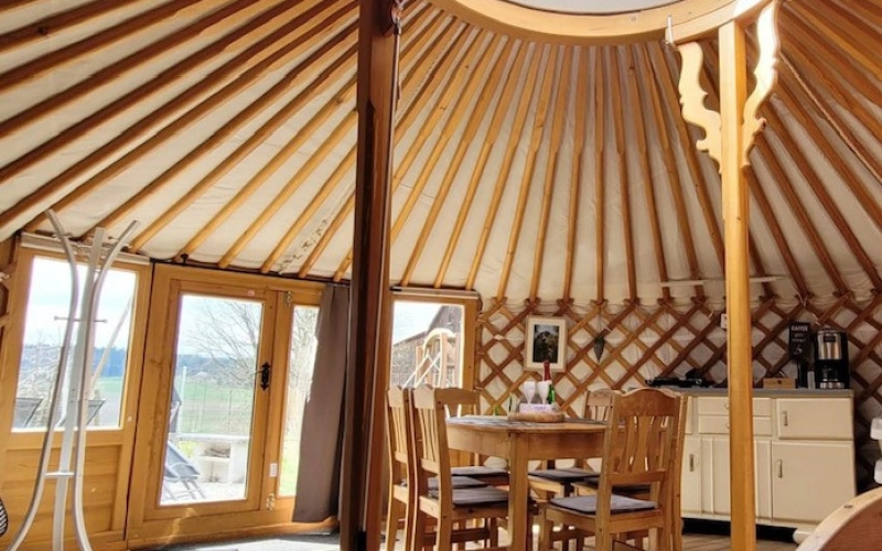 Blick in eine eingerichtete Jurte: Holzgitterwand, Speichendach und in der Mitte die runde Lichtkuppel, darunter Esstisch, K&uuml;chenzeile und Bett.