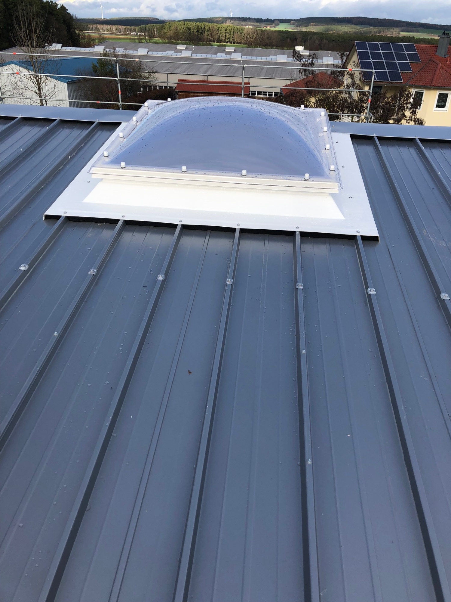 Die fertige Lichtkuppel auf dem grauen Trapezblechdach, dahinter Hallen, Wohnhäuser und bewaldete Hügel.
