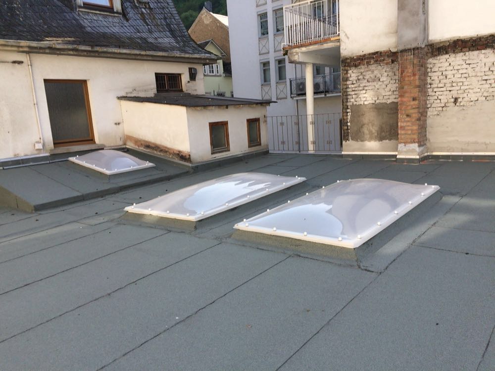 Bitumen-Flachdach im Hinterhof mit drei neu montierten Lichtkuppeln unterschiedlicher Größe, dahinter Wohnhäuser mit Balkonen.