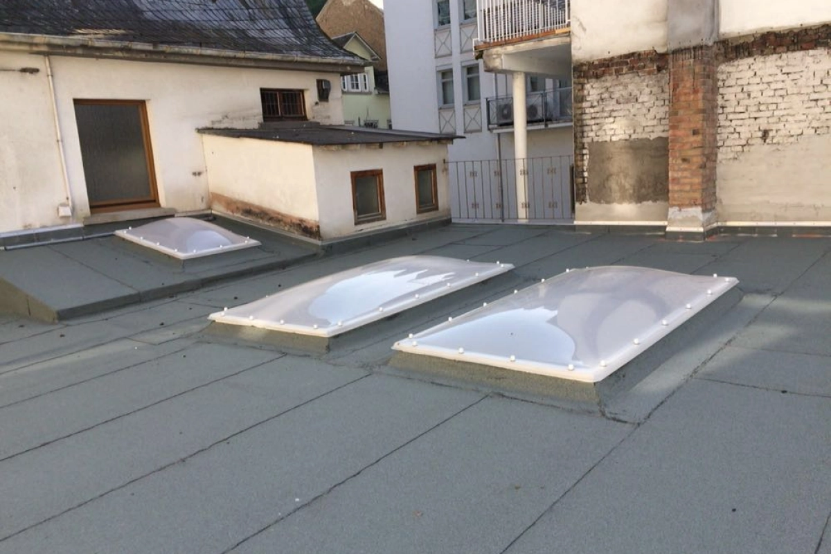 Flachdach mit drei rechteckigen Lichtkuppeln auf wei&szlig;en Aufsatzkr&auml;nzen, dahinter Wohnh&auml;user mit Balkonen.