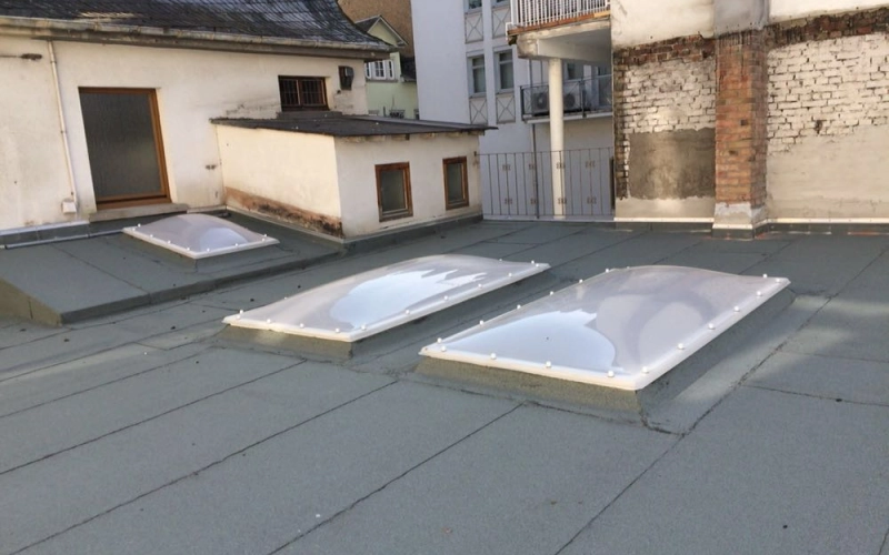Flachdach mit drei rechteckigen Lichtkuppeln auf wei&szlig;en Aufsatzkr&auml;nzen, dahinter Wohnh&auml;user mit Balkonen.