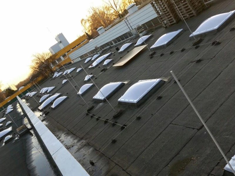 Schr&auml;ger Blick &uuml;ber das Dach mit neuen Lichtkuppeln in mehreren Reihen, im Hintergrund eine gelbe Fassade und gestapelte Paletten.