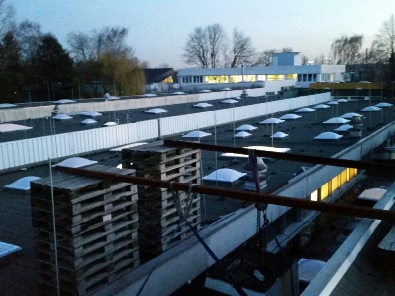 Dachfl&auml;che in der D&auml;mmerung mit gestapelten Europaletten, dahinter erleuchtete B&uuml;rofenster und viele Lichtkuppeln.