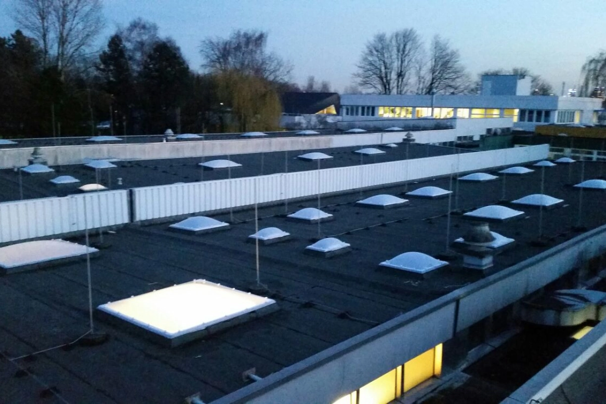Dachfl&auml;chen mit Lichtkuppeln in der D&auml;mmerung, im Hintergrund ein Geb&auml;ude mit erleuchteten Fenstern.