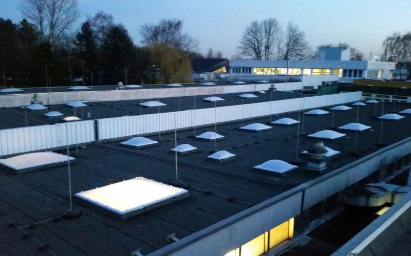 Dachfl&auml;chen mit Lichtkuppeln in der D&auml;mmerung, im Hintergrund ein Geb&auml;ude mit erleuchteten Fenstern.