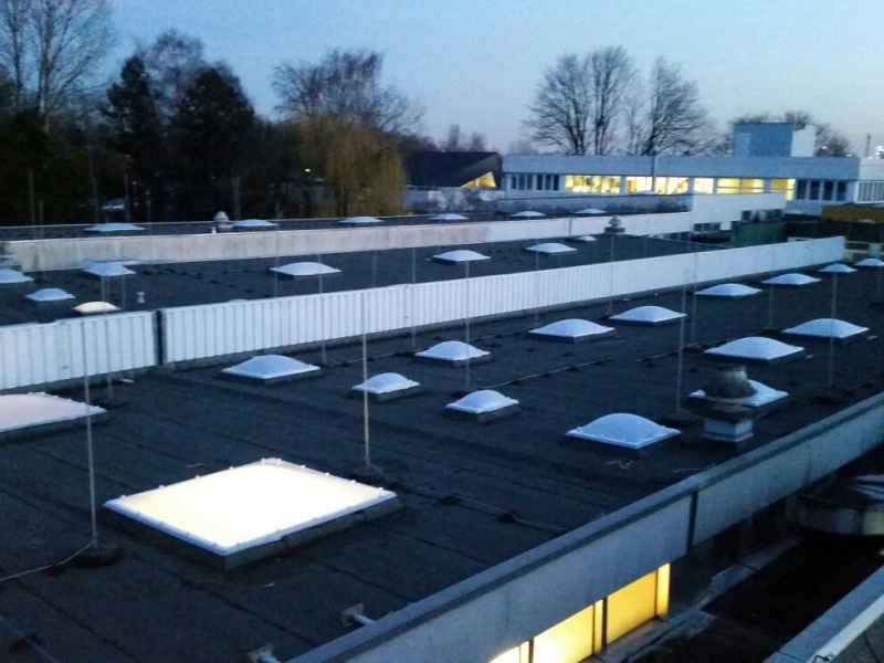 Dachfl&auml;chen mit Lichtkuppeln in der D&auml;mmerung, im Hintergrund ein Geb&auml;ude mit erleuchteten Fenstern.