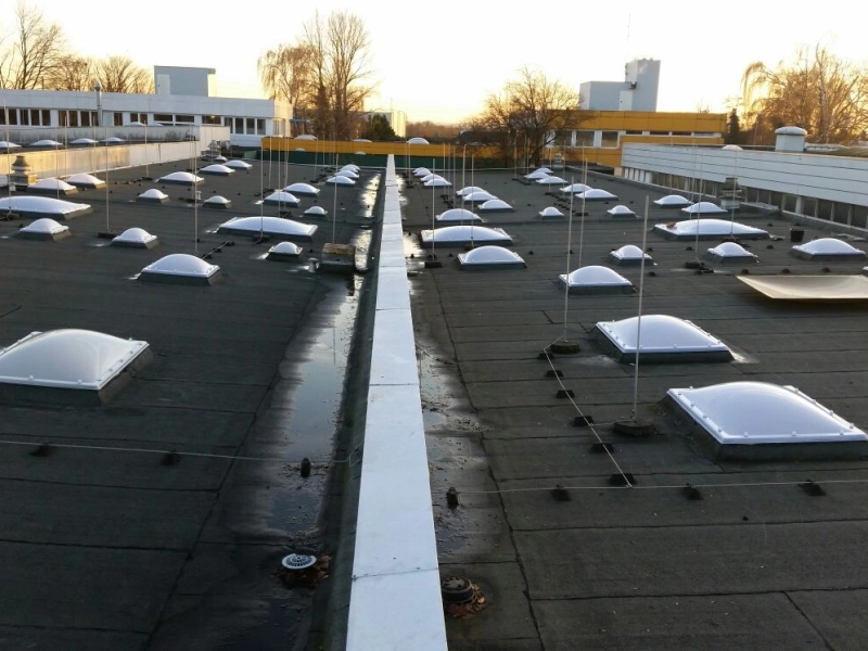 Zwei Dachh&auml;lften voller neuer Lichtkuppeln, mittig eine wei&szlig;e Attika, im Hintergrund B&uuml;rotrakt und gelbe Fassade.