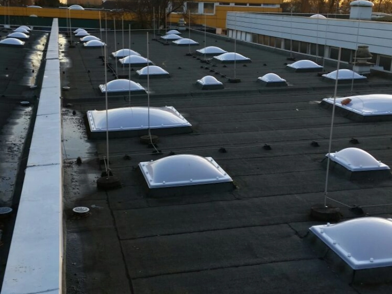 Hochformatiger Blick &uuml;ber die Dachfl&auml;che mit zwei Reihen neuer Lichtkuppeln, dahinter ein Flachbau im Abendlicht.