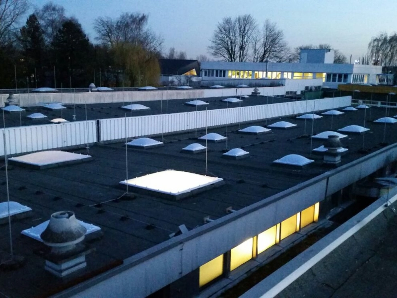 Dachfl&auml;che in der D&auml;mmerung mit vielen Lichtkuppeln, zwei davon von innen beleuchtet, dahinter ein beleuchteter B&uuml;rotrakt.