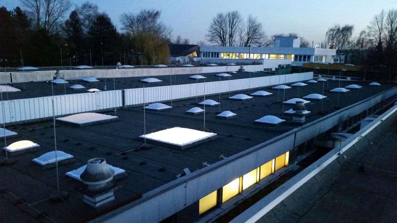 Dachfläche in der Dämmerung mit vielen Lichtkuppeln, zwei davon von innen beleuchtet, dahinter ein beleuchteter Bürotrakt.