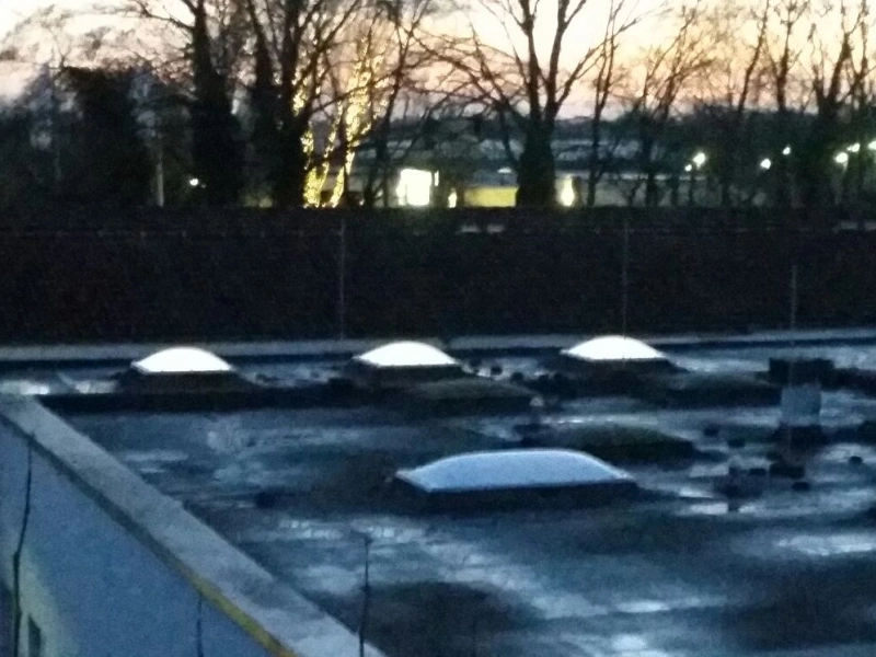 Dachfl&auml;che bei Nacht mit von innen beleuchteten Lichtkuppeln, dahinter eine Hecke und Stra&szlig;enlaternen.