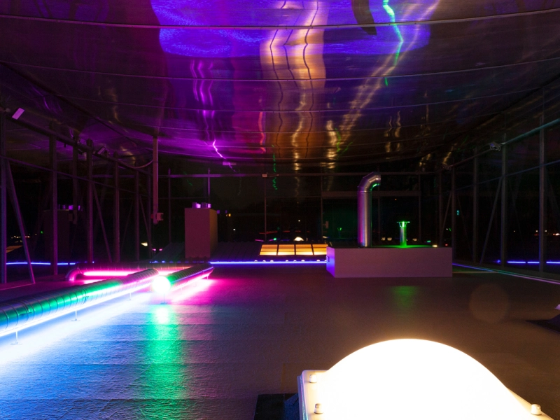 Weitwinkelblick durch den Pavillon: farbig gefasste Rohre auf der linken Seite, rechts zwei leuchtende Lichtkuppeln.