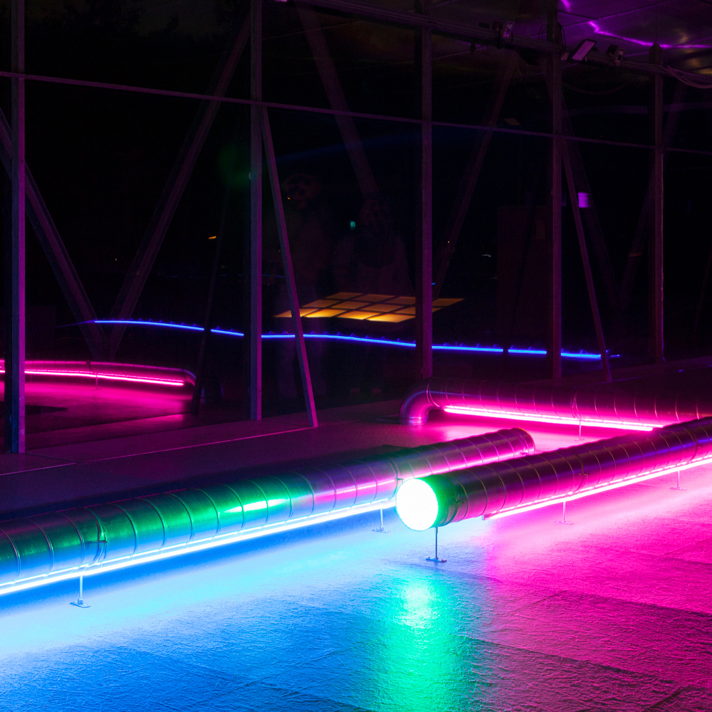 Nahaufnahme der Installation: Lüftungsrohre, an denen entlang blaue, grüne und pinke Leuchtstreifen verlaufen.